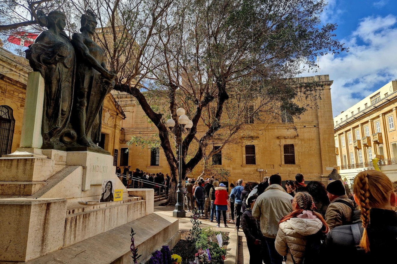Hosszú sor a Szent János-társkatedrális előtt. A Nagy Ostrom-emlékműn Daphne Caruana Galizia oknyomozó újságíró fényképe, aki korrupciós ügyeket tárt fel, 2017-ben meggyilkolták, a tettest azóta is „keresik”