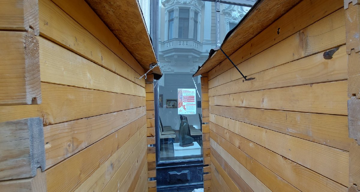 A bódék mögött egy galéria kirakata sejlik elő. Ismétlem: galéria. Képzőművészet. Ugye, olyan valami, amit elsősorban látni, nézni kell. Kellene. Ha lehetne. 