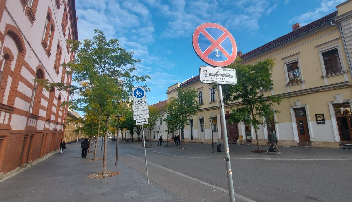 A tábla egyértelműen jelzi: itt megállni tilos. A másik pedig azt, hogy ez egy gyalogos övezet. Kivéve iskolabuszok, biciklik, illetve intézményi, vállalati ellátás. Pedig nem gyalogos övezet, hanem shared space. Ahol az autók is járhatnak. Ja, és az ottlakókról sem esik szó. Riveran, ez a román kifejezés az ottlakókra. Ez lenne, ha ott lenne a táblán.
